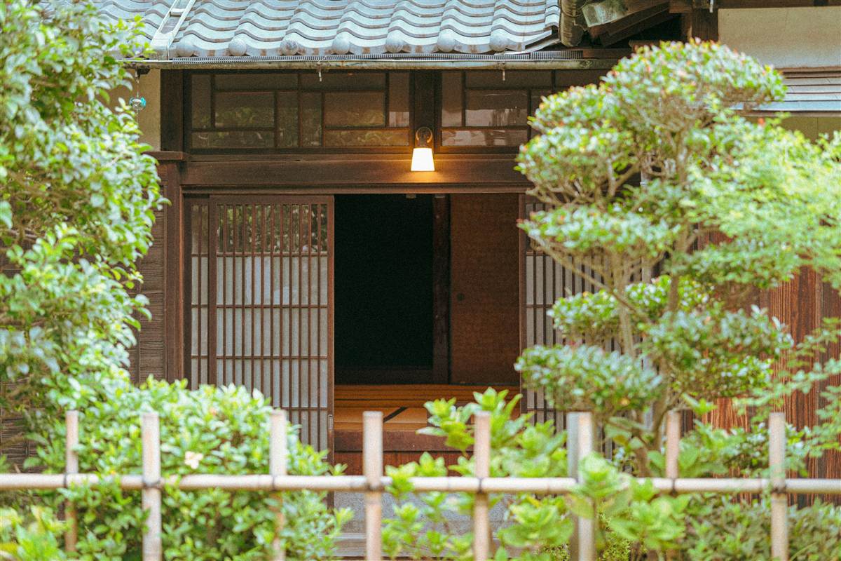 植木に囲まれた古民家の玄関と、ともった灯り（古民家カフェ●●のサンプルサイトより）