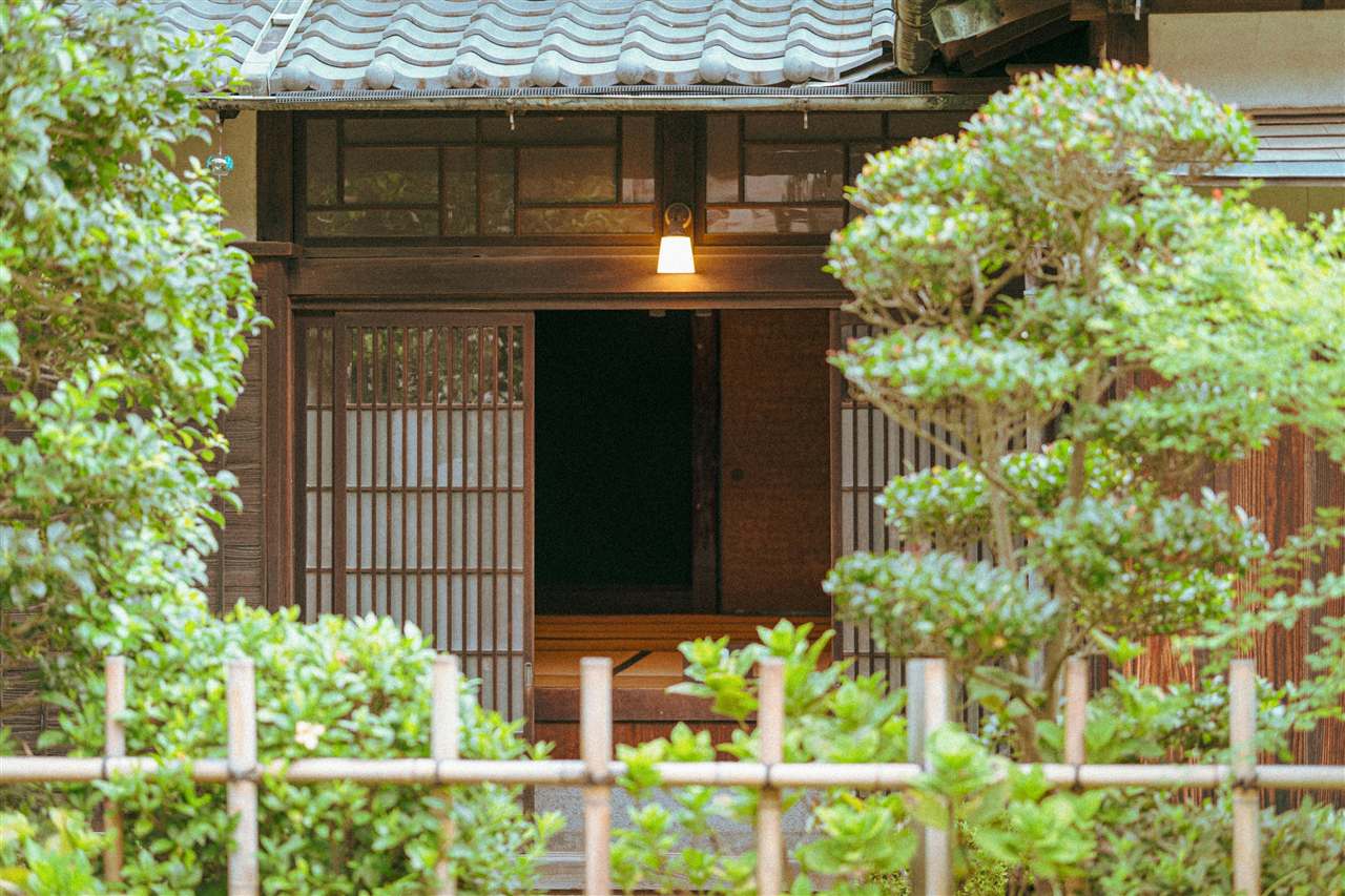植木に囲まれた古民家の玄関と、ともった灯り（Unsplashの写真）